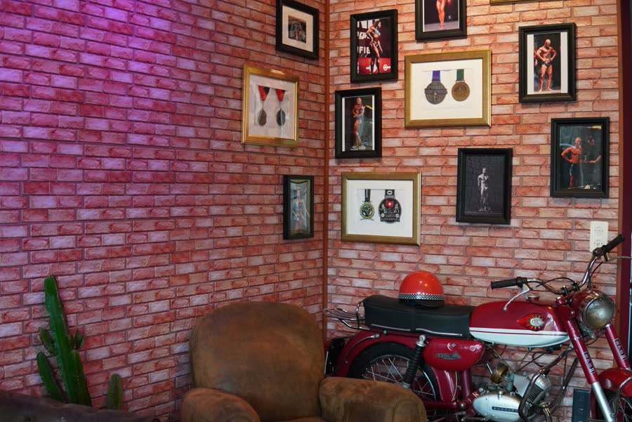 Coin lounge à l'entrée : mur de briques rouges, médailles et photos de compétition encadrées, fauteuil en cuir et moto ancienne